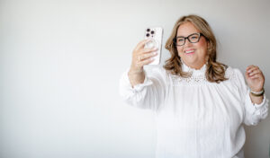 Photo of a white woman with blonde hair and glasses looking at her phone, taking a selfie, smiling on a white background.